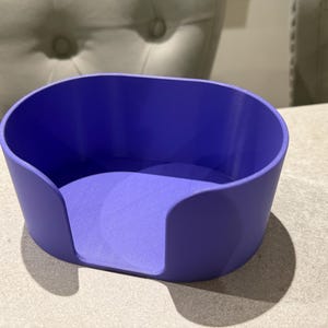 May include: A solid purple, oval-shaped container with rounded sides and a flat base. The container has a smooth, matte finish and is designed to hold small items. The object is sitting on a light-colored surface.
