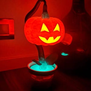 May include: A glowing orange pumpkin-shaped lamp with a carved face, set in a small pot with a blue glow. The lamp is on a dark surface, with a red-toned background. A decorative Halloween-themed item.