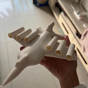 May include: A white airplane-shaped object with cylindrical tubes on the wings. The toy plane has a pointed nose and a sleek design. The object is held in a person's hand.