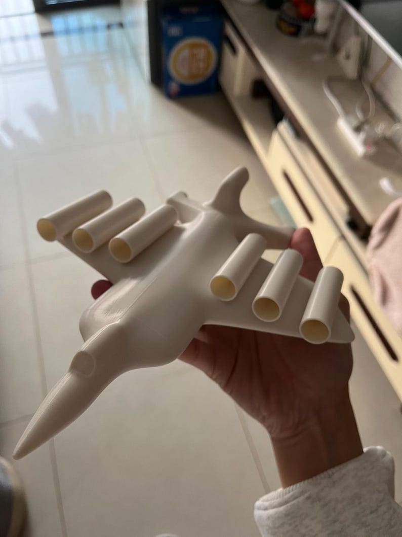 May include: A white airplane-shaped object with cylindrical tubes on the wings. The toy plane has a pointed nose and a sleek design. The object is held in a person's hand.