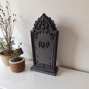 May include: A dark gray tombstone with the letters "RIP" carved into the front. The top of the tombstone features skull and floral detailing. The tombstone is a decorative item, likely for Halloween or gothic decor.