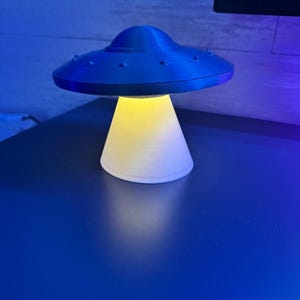 May include: A blue UFO-shaped lamp with a bright yellow light emanating from its base. The lamp is made of plastic and sits on a blue surface. The lamp's design is reminiscent of a classic flying saucer.