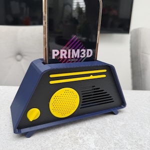 May include: A navy blue and black phone stand with a yellow speaker and accents. The stand holds a phone with the text "PRIM3D" displayed on the screen. The stand has a retro, geometric design.