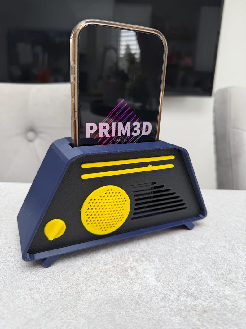 May include: A navy blue and black phone stand with a yellow speaker and accents. The stand holds a phone with the text "PRIM3D" displayed on the screen. The stand has a retro, geometric design.