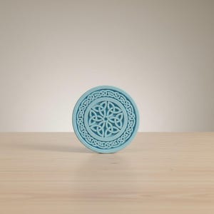 May include: A round, light blue coaster with a Celtic knot design. The coaster features a central knot pattern surrounded by a circular border of interwoven knots. The coaster is made of a solid material and is suitable for holding beverages.