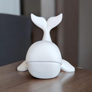 May include: A white whale figurine with a textured surface. The whale has a rounded body, flipper-like appendages, and a prominent tail fin. The figurine is placed on a wooden surface.