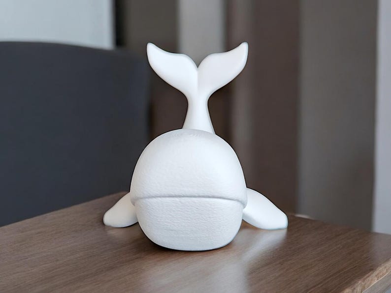May include: A white whale figurine with a textured surface. The whale has a rounded body, flipper-like appendages, and a prominent tail fin. The figurine is placed on a wooden surface.