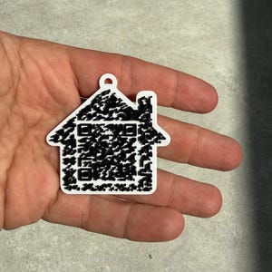 May include: A white house-shaped keychain with a black QR code design. The keychain has a small loop at the top for attachment. The house shape includes a roof, windows, and a chimney.