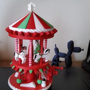 Op de afbeelding: Een rood-witte carrousel met witte paarden, een groene kerstboom en een sneeuwpopfiguur. De carrousel heeft een gestreept rood, wit en groen dak en een witte sneeuwvlok bovenop. Op de achtergrond staat een blauwe ballonhond.