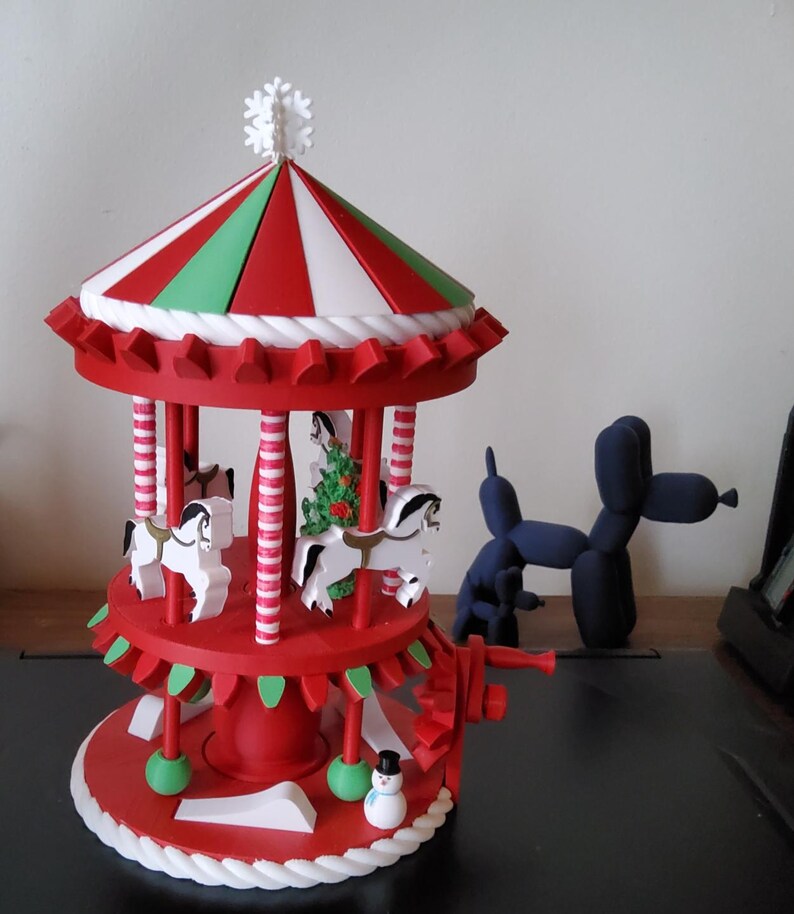 Op de afbeelding: Een rood-witte carrousel met witte paarden, een groene kerstboom en een sneeuwpopfiguur. De carrousel heeft een gestreept rood, wit en groen dak en een witte sneeuwvlok bovenop. Op de achtergrond staat een blauwe ballonhond.