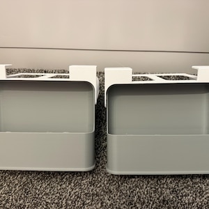 May include: Two gray, rectangular storage bins with white over-the-door hooks. The bins are made of a solid material and are designed to hang over a door. The bins are empty and sit on a gray carpet.