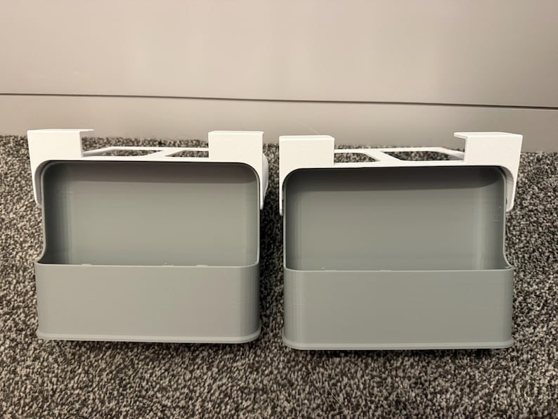 May include: Two gray, rectangular storage bins with white over-the-door hooks. The bins are made of a solid material and are designed to hang over a door. The bins are empty and sit on a gray carpet.