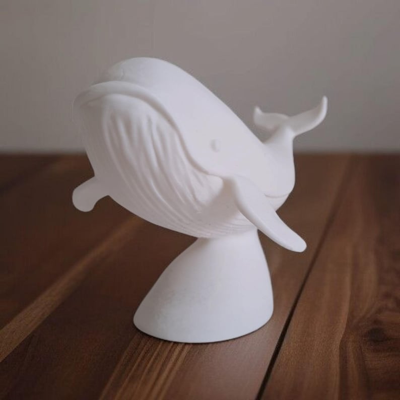 K&ouml;nnte beinhalten: Eine wei&szlig;e Wal-Skulptur mit glatter, matter Oberfl&auml;che. Der Wal ist in einer dynamischen Pose dargestellt, mit gebogenem K&ouml;rper und ausgestreckten Flossen. Die Skulptur ist auf einem abgerundeten Sockel montiert.