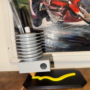 May include: A gray pen holder with a unique design, featuring a series of stacked, circular tiers. The holder has a rectangular base and a yellow, curved accent piece. Two pens are visible inside the holder. The background includes a painting of a motorcycle.