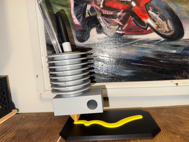 May include: A gray pen holder with a unique design, featuring a series of stacked, circular tiers. The holder has a rectangular base and a yellow, curved accent piece. Two pens are visible inside the holder. The background includes a painting of a motorcycle.