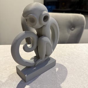 Puede incluir: Una escultura de mono gris con una cola rizada y grandes rasgos similares a orejas. El mono est&aacute; sentado sobre una base rectangular. La escultura est&aacute; hecha de un material s&oacute;lido y est&aacute; pintada con un acabado gris mate. Es un art&iacute;culo decorativo.