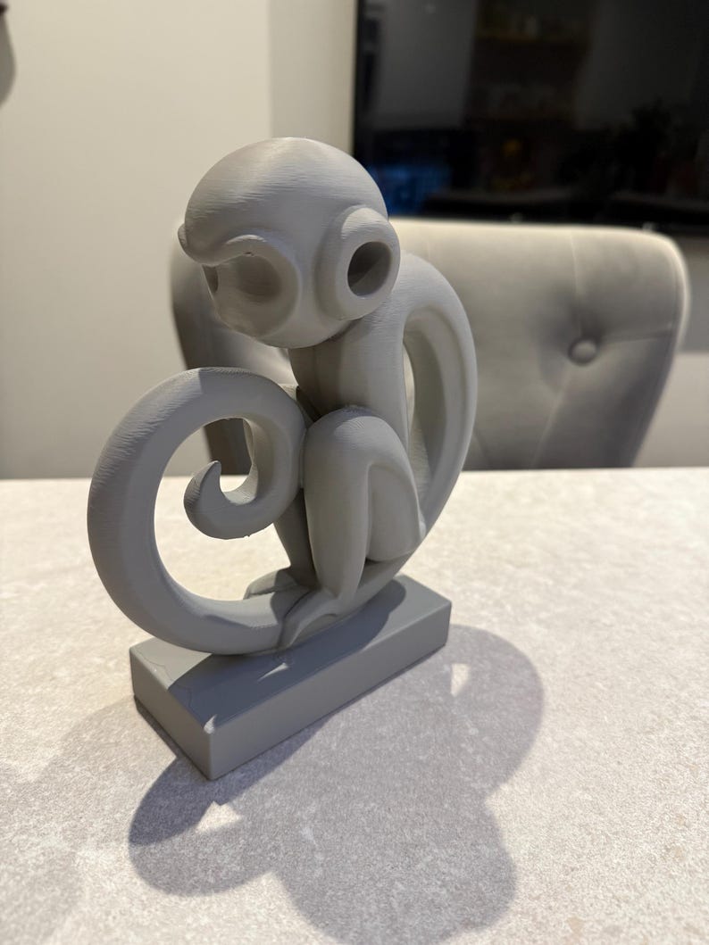Puede incluir: Una escultura de mono gris con una cola rizada y grandes rasgos similares a orejas. El mono est&aacute; sentado sobre una base rectangular. La escultura est&aacute; hecha de un material s&oacute;lido y est&aacute; pintada con un acabado gris mate. Es un art&iacute;culo decorativo.