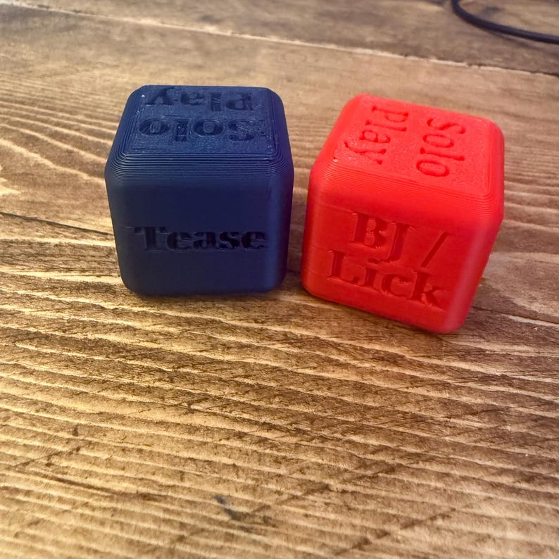 Puede incluir: Dos dados impresos en 3D, uno azul y otro rojo, est&aacute;n sobre una superficie de madera. El dado azul tiene las palabras "Tease" y "Solo Play" en sus lados. El dado rojo tiene "BJ / Lick" y "Solo Play".