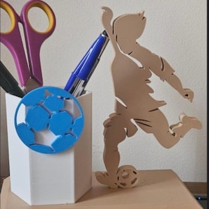 May include: A desk organizer featuring a soccer theme. It includes a white pen holder with purple scissors, a blue pen, and a blue soccer ball cutout. A wooden cutout of a soccer player kicking a ball is also present.