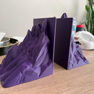 May include: A pair of purple mountain-shaped bookends. The bookends have a textured, rocky appearance and a flat back. They are designed to hold books upright on a desk or shelf. The bookends are made of a solid material.