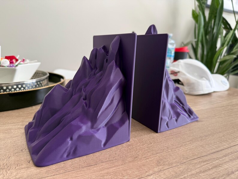 May include: A pair of purple mountain-shaped bookends. The bookends have a textured, rocky appearance and a flat back. They are designed to hold books upright on a desk or shelf. The bookends are made of a solid material.