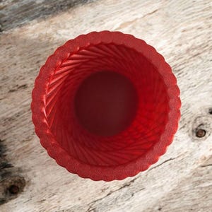 May include: A top-down view of a red, textured vase. The vase has a spiral design on the inside and a scalloped edge. The vase is made of a solid red material.