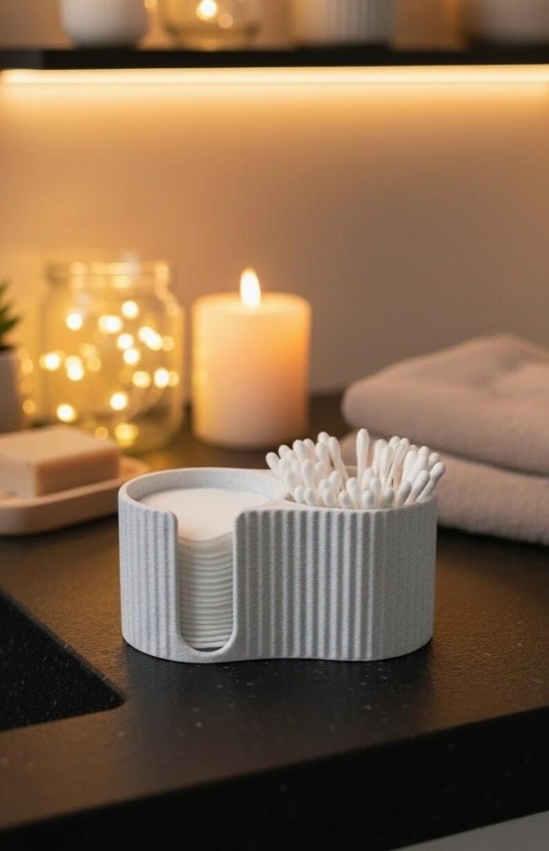 May include: A light gray, ribbed, two-compartment bathroom organizer. One side holds cotton pads, the other holds cotton swabs. A lit candle and a jar of fairy lights are in the background, suggesting a spa-like setting.