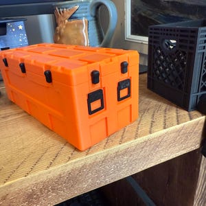 May include: Bright orange storage container with black latches and handles. The rectangular box has a textured surface and is sitting on a wooden surface. A black mouse and a blue box are in the background.