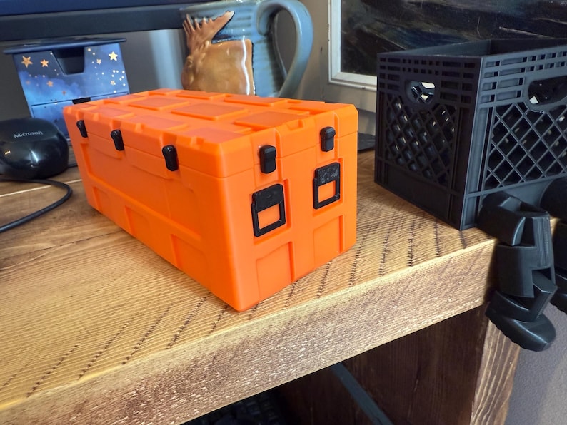 May include: Bright orange storage container with black latches and handles. The rectangular box has a textured surface and is sitting on a wooden surface. A black mouse and a blue box are in the background.