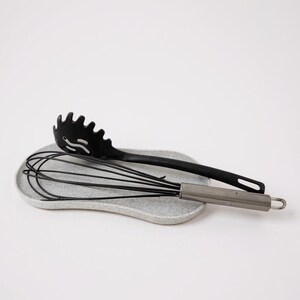 May include: A black spaghetti server and a stainless steel whisk rest on a speckled grey ceramic spoon rest. The kitchen tools are arranged on a white background. The spaghetti server has a slotted design.