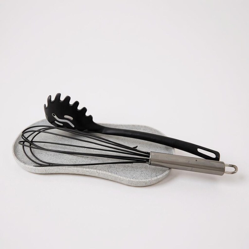 May include: A black spaghetti server and a stainless steel whisk rest on a speckled grey ceramic spoon rest. The kitchen tools are arranged on a white background. The spaghetti server has a slotted design.