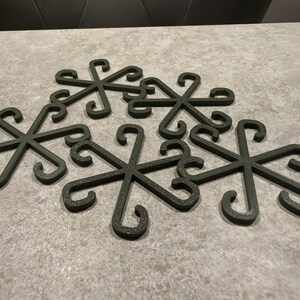 May include: Six dark green metal trivets arranged on a gray surface. Each trivet has a snowflake-like design with curved hooks at the ends of each spoke. The trivets are made of a textured material.