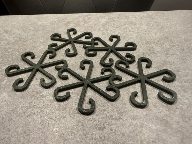 May include: Six dark green metal trivets arranged on a gray surface. Each trivet has a snowflake-like design with curved hooks at the ends of each spoke. The trivets are made of a textured material.