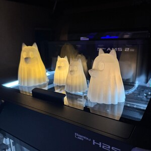 Pu&ograve; includere: Quattro figurine bianche traslucide a forma di cane con interni luminosi sono esposte su una superficie nera. Le figurine sono illuminate dall'interno, creando un bagliore morbido e caldo. La base del display riporta la scritta "Bambu Lab H2S".