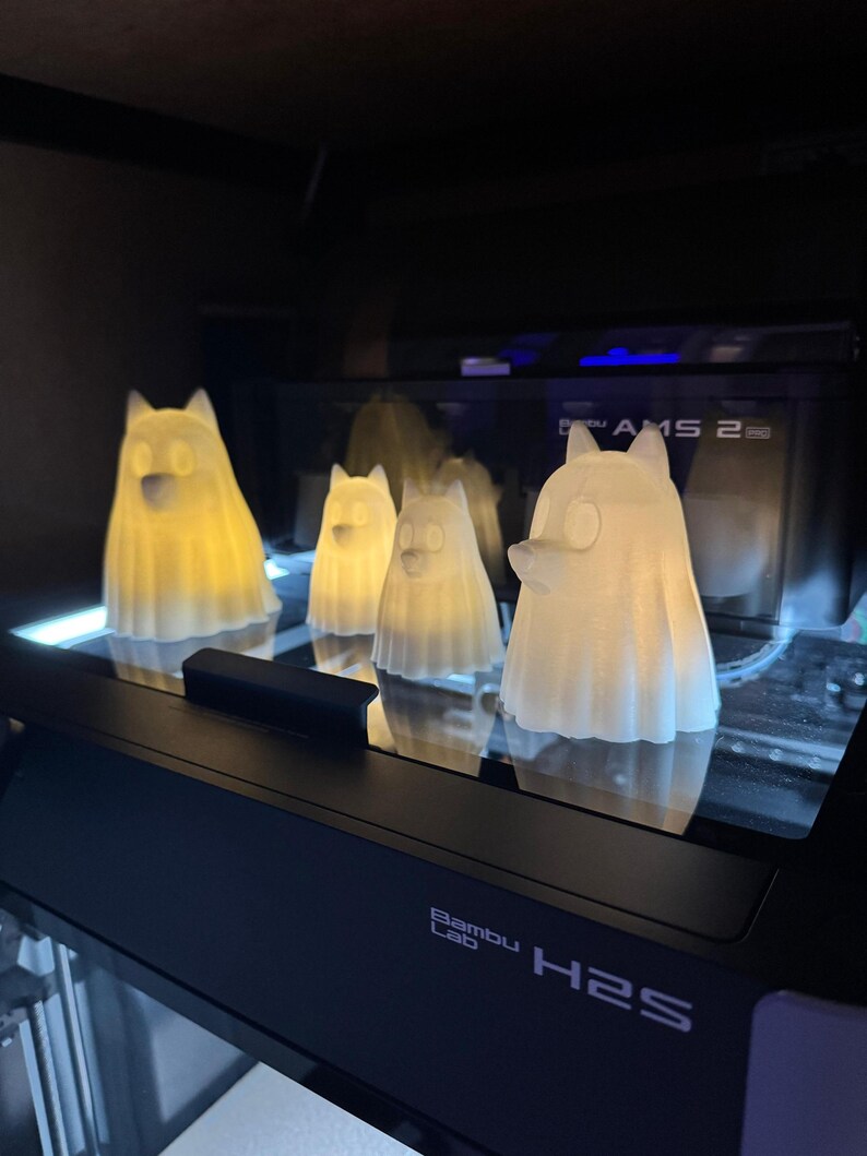Pu&ograve; includere: Quattro figurine bianche traslucide a forma di cane con interni luminosi sono esposte su una superficie nera. Le figurine sono illuminate dall'interno, creando un bagliore morbido e caldo. La base del display riporta la scritta "Bambu Lab H2S".