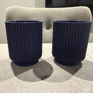 May include: Two navy blue cylindrical planters with vertical ribbed detailing. The planters are made of a solid material and have a rounded base. They are sitting on a light gray surface, and the shadows are visible.