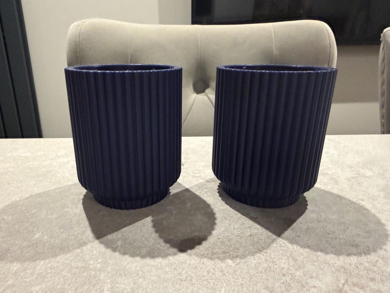 May include: Two navy blue cylindrical planters with vertical ribbed detailing. The planters are made of a solid material and have a rounded base. They are sitting on a light gray surface, and the shadows are visible.