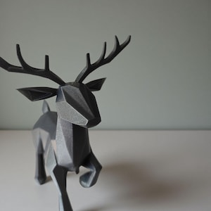 May include: A dark gray geometric deer sculpture with a faceted design. The deer has large antlers and is posed in a running stance. The sculpture is made of a solid material and is set against a neutral background.