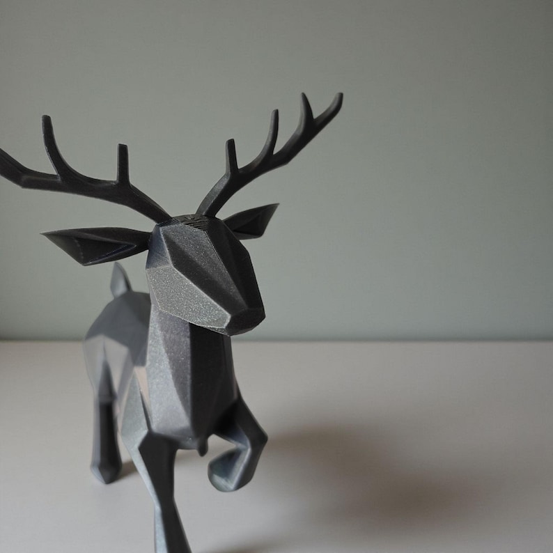 May include: A dark gray geometric deer sculpture with a faceted design. The deer has large antlers and is posed in a running stance. The sculpture is made of a solid material and is set against a neutral background.