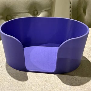 May include: A solid purple, oval-shaped container with curved sides and a cutout on one side. The container sits on a light-colored surface, casting a shadow. The background includes two upholstered chairs.