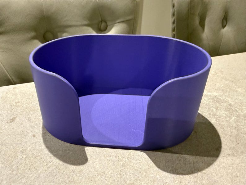 May include: A solid purple, oval-shaped container with curved sides and a cutout on one side. The container sits on a light-colored surface, casting a shadow. The background includes two upholstered chairs.