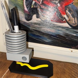 May include: A gray and black desk organizer with a pen holder, a small rectangular compartment, and a yellow decorative element. The pen holder contains a silver pen and a black marker. The organizer sits on a wooden surface, with a painting of a motorcycle in the background.