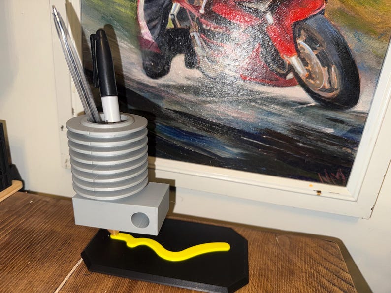 May include: A gray and black desk organizer with a pen holder, a small rectangular compartment, and a yellow decorative element. The pen holder contains a silver pen and a black marker. The organizer sits on a wooden surface, with a painting of a motorcycle in the background.