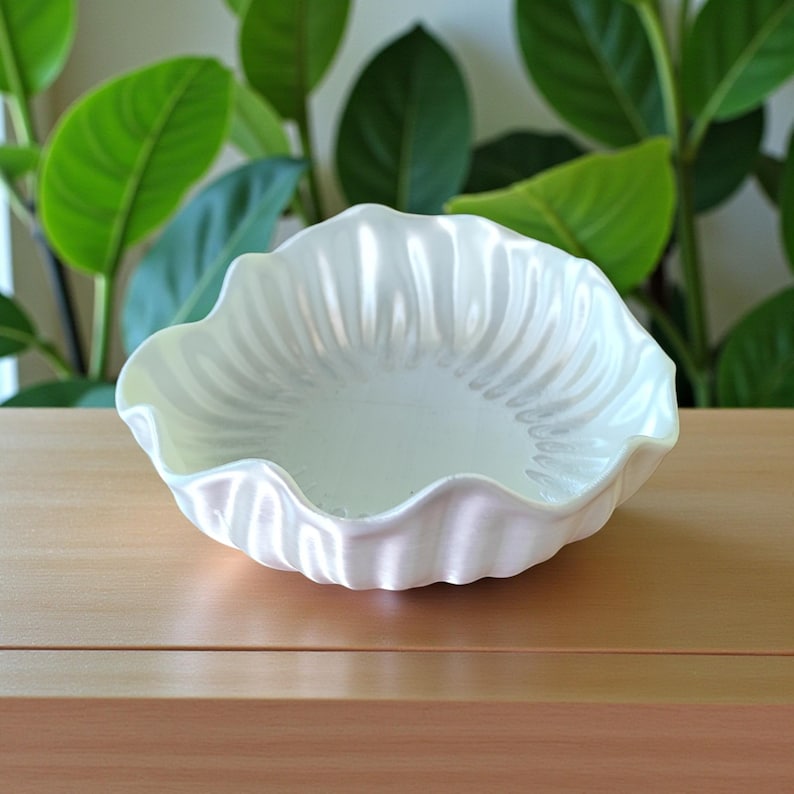 May include: A white, scalloped-edge bowl with a pearlescent finish. The bowl has a textured interior and sits on a wooden surface. Green leaves are visible in the background. This decorative bowl is ideal for serving or display.
