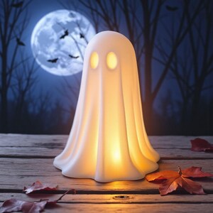 May include: A glowing white ghost-shaped lamp with two oval eye cutouts. The lamp is on a wooden surface with fallen leaves, set against a backdrop of a full moon, bats, and silhouetted trees. A Halloween decoration.