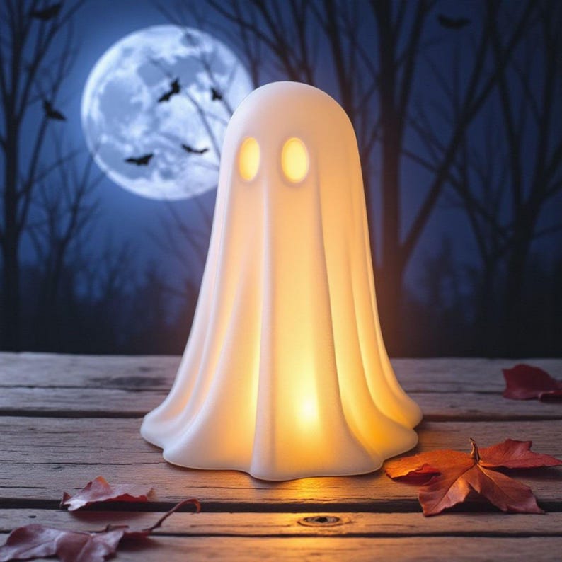 May include: A glowing white ghost-shaped lamp with two oval eye cutouts. The lamp is on a wooden surface with fallen leaves, set against a backdrop of a full moon, bats, and silhouetted trees. A Halloween decoration.