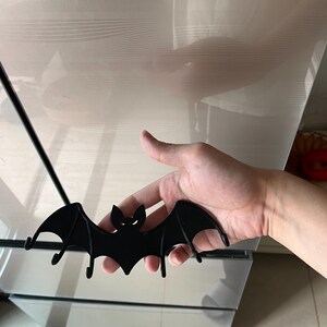 May include: A black bat-shaped wall hook with a minimalist design. The bat has outstretched wings and two small hooks underneath. The eyes and mouth are cut out, creating a spooky Halloween decoration.