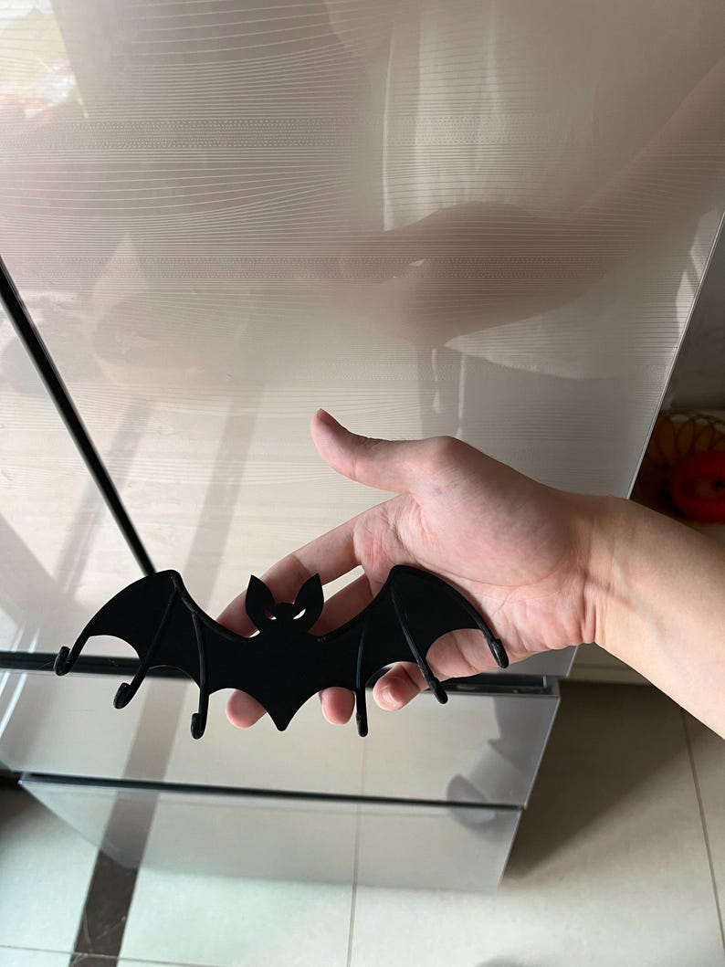 May include: A black bat-shaped wall hook with a minimalist design. The bat has outstretched wings and two small hooks underneath. The eyes and mouth are cut out, creating a spooky Halloween decoration.