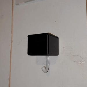 May include: A black, square-shaped wall hook with a curved, clear plastic hook extending downwards. The hook is mounted on a white wall, providing a simple and functional storage solution. The design is minimalist and modern.