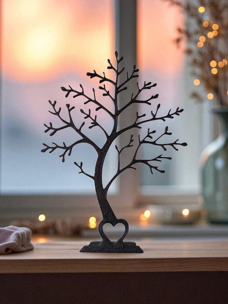May include: A black metal tree-shaped jewelry holder with a heart-shaped base. The tree has many branches for hanging necklaces and earrings. The background shows a window with a sunset and a vase with decorative lights.